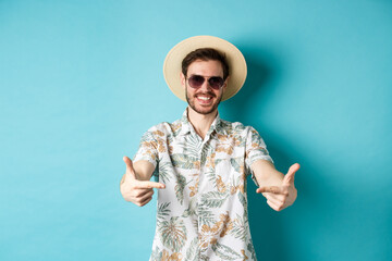 Happy tourist in summer hat and hawaiian shirt, pointing fingers at logo on center, showing something, standing on blue background