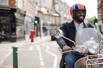 Smiling young businessman in helmet riding motor scooter on urban street