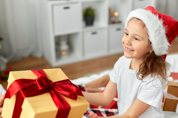 christmas, holidays and childhood concept - happy girl in santa hat with gift box at home