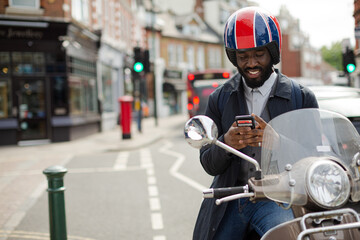 Smiling young businessman in helmet on motor scooter texting with cell phone on urban street