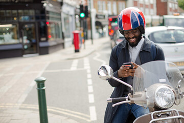 Smiling young businessman in helmet on motor scooter texting with cell phone on urban street