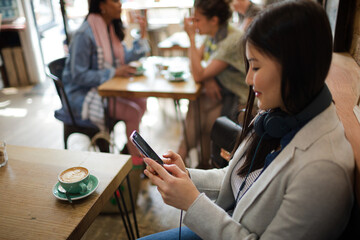 Young woman with headphones texting with cell phone and drinking coffee at cafe table