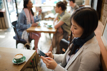 Young woman with headphones texting with cell phone and drinking coffee at cafe table
