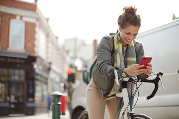 Obraz premium Young woman commuting on bicycle, texting with cell phone on sunny urban street