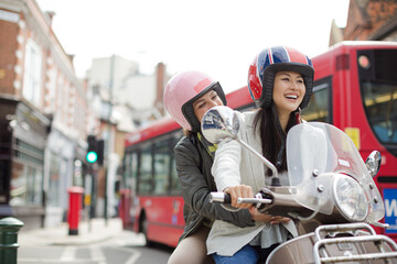 Obraz premium Smiling young women friends wearing helmets, riding motor scooter on urban street