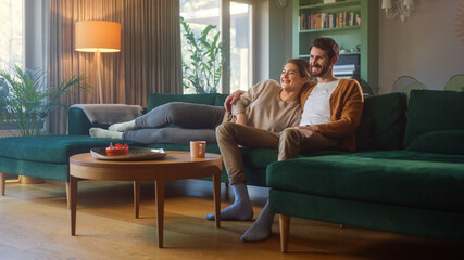 Couple Watches TV together while Sitting on a Couch in the Living Room. Girlfriend and Boyfriend embrace, cuddle, talk, smile and watch Television Streaming Services. Home with Cozy Stylish Interior. - Powered by Adobe