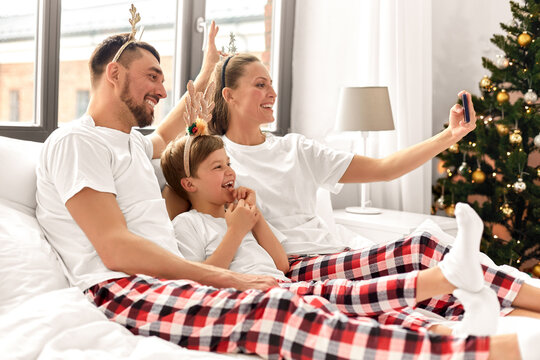 Family, Winter Holidays And People Concept - Happy Mother, Father And Little Son In Matching Pajamas Taking Selfie With Smartphone In Bed In Christmas Morning