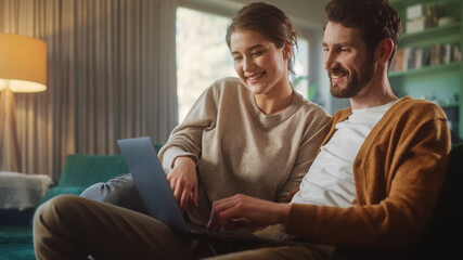 Couple Use Laptop Computer, while Sitting on a Couch in the Apartment. Boyfriend and Girlfriend Talk, Shop on Internet, Choose Product to Order Online, Watch Funny Videos on Streaming Service