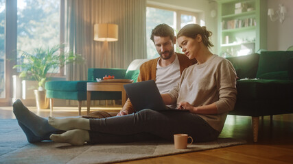 Couple Use Laptop Computer, while Sitting on the Living Floor room of their Apartment. Boyfriend and Girlfriend Talk, Shop on Internet, Choose Product to Order Online, Doing Remote Work Together