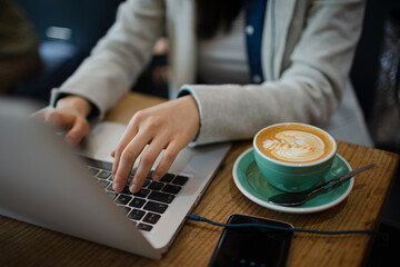 Hands of young woman using laptop, drinking cappuccino in cafe