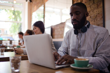 Man with headphones using laptop and drinking coffee in cafe