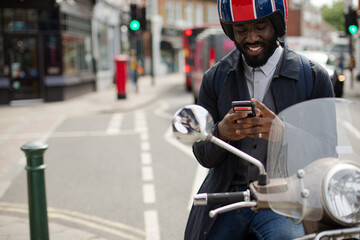 Smiling young businessman in helmet on motor scooter texting with cell phone on urban street
