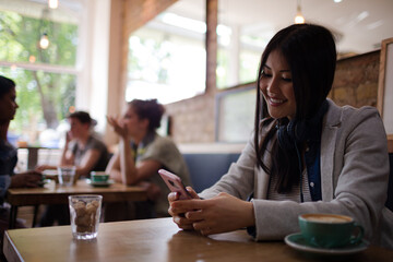 Young woman with headphones texting with cell phone and drinking coffee at cafe table