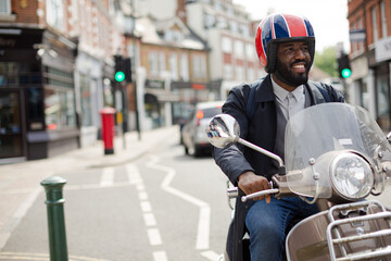Fototapeta premium Smiling young businessman in helmet riding motor scooter on urban street