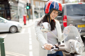 Smiling young woman texting with cell phone on motor scooter, wearing helmet on urban street