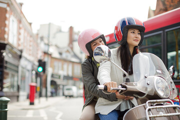 Fototapeta na wymiar Smiling young women friends wearing helmets, riding motor scooter on urban street