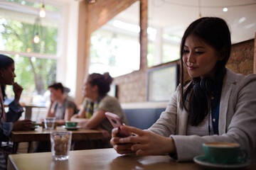 Young woman with headphones texting with cell phone and drinking coffee at cafe table