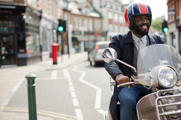 Smiling young businessman in helmet riding motor scooter on urban street