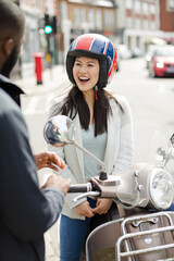 Fototapeta na wymiar Smiling young woman in helmet on motor scooter, talking to friend on urban street
