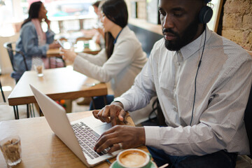 Man with headphones using laptop and drinking coffee in cafe