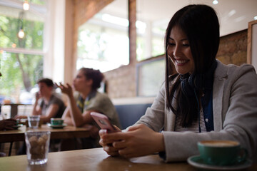 Young woman with headphones texting with cell phone and drinking coffee at cafe table