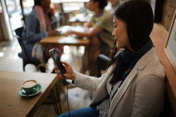 Young woman with headphones texting with cell phone and drinking coffee at cafe table