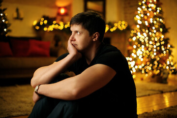 Lonely young man bored in a room decorated with a festive christmas tree and garlands on new years...