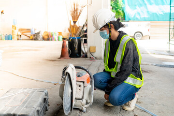 Young Asian female engineer wearing a medical mask to cover her mouth and wearing a white safety helmet A small concrete slab cutting machine is being prepared in the construction Zone.