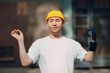 Disabled european man with artificial limb of hand meditating outdoors. Handsome young guy wears t-shirt and hat enjoys sunny day. Concept of mental health. Modern medical technology today