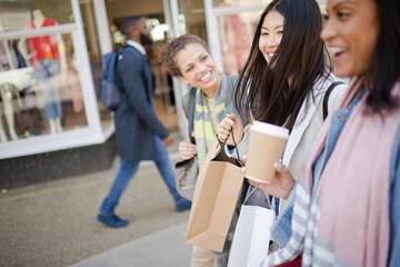 Laughing women friends walking along storefront with coffee and shopping bags