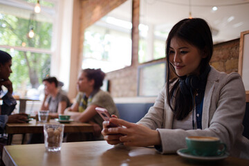 Young woman with headphones texting with cell phone and drinking coffee at cafe table