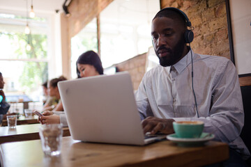 Man with headphones using laptop and drinking coffee in cafe