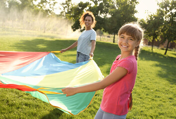 Cute child and African-American teacher playing with rainbow playground parachute on green grass....