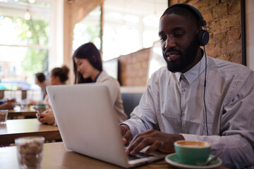 Man with headphones using laptop and drinking coffee in cafe
