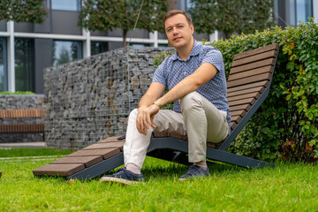 Obraz premium Portrait of smiling handsome millennial man. Positive mature man looking at camera, outdoor backyard at office building recreational area. Handsome man relaxing on wooden chaise lounge in city park.