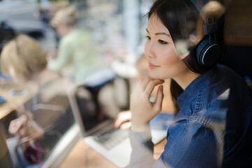 Pensive young woman listening to music with headphones looking away at cafe window
