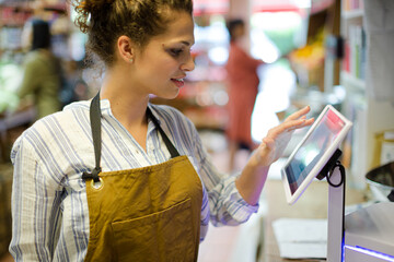Female cashier using touch screen cash register in grocery store - obrazy, fototapety, plakaty
