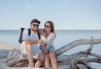 young attractive smiling happy man and woman in sunglasses sitting on log at sand beach.