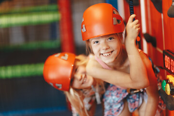 Two happy little girls in red helmets climbing the wall in bouldering center. School girls smiling at the camera and having fun in indoor playground for children. Indoor climbing class for kids