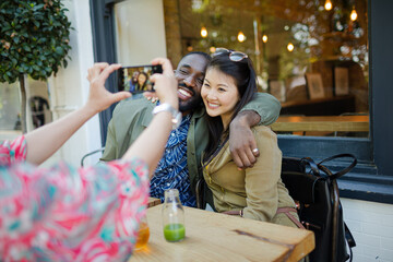 Woman photographing affectionate couple friends with camera phone at sidewalk cafe
