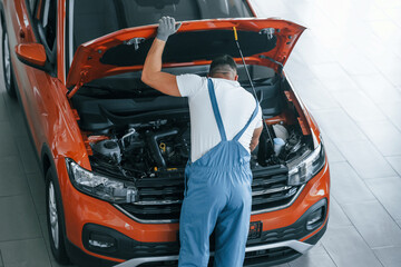 In blue uniform. Man is repairing broken automobile indoors