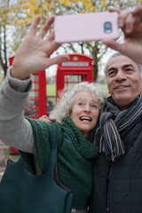 Smiling senior couple taking selfie in autumn park in front of red telephone booths