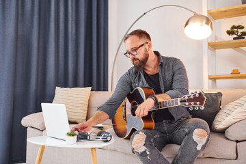 On the sofa. Playing acoustic guitar. Man in casual clothes is indoors