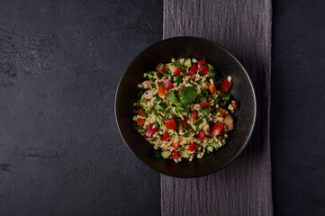 Traditional salad tabbouleh made of bulgur or couscous, poultry meat, parsley, mint in dark bowl with napkin on black background