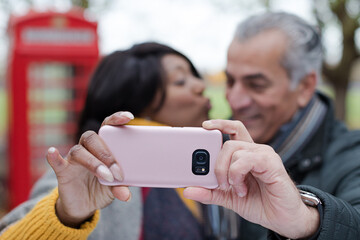 Smiling senior couple taking selfie in autumn park in front of red telephone booths