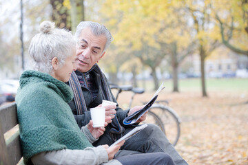Senior couple reading newspaper and drinking coffee on bench in autumn park