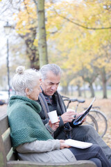 Senior couple reading newspaper and drinking coffee on bench in autumn park