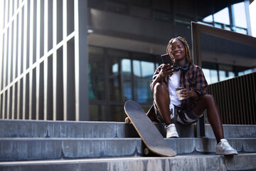 Fashion African woman with skateboard. Young stylish woman with skateboard using the phone outdoors