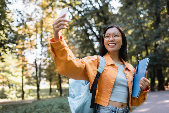 cheerful asian woman with backpack and notebooks taking selfie on cellphone in park