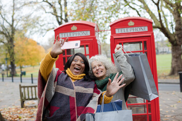 Senior women friends with shopping bags taking selfie in autumn park in front of red telephone booths
